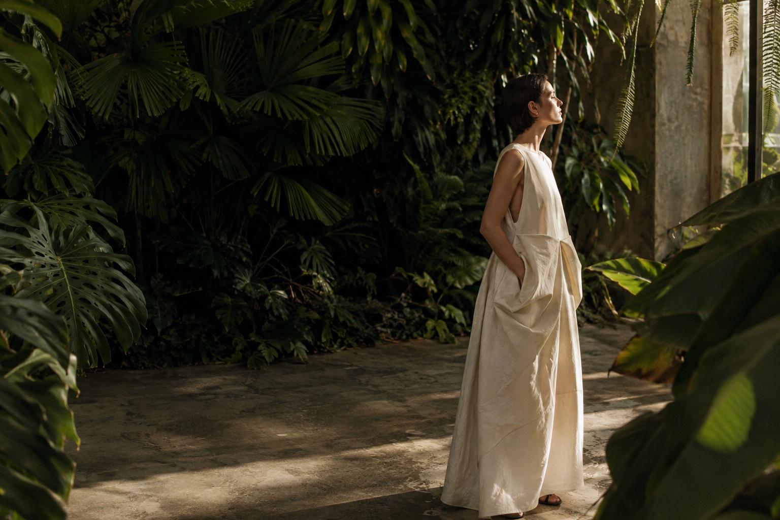 An editorial portrait in a sunlit greenhouse, with an ivory dress framed by deep green tropical leaves